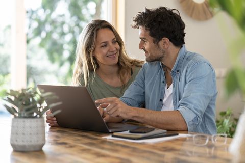 smiling couple looking at each other while sitting in front of laptop at table