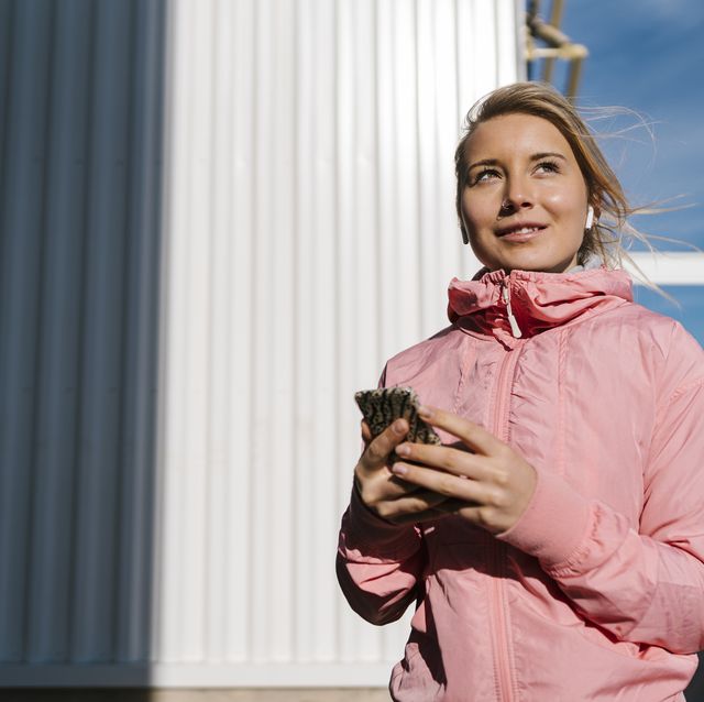smiling female athlete with in ear headphones looking away while holding mobile phone while against wall