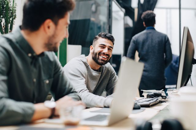 smiling male colleagues looking at each other while sitting by desk in office