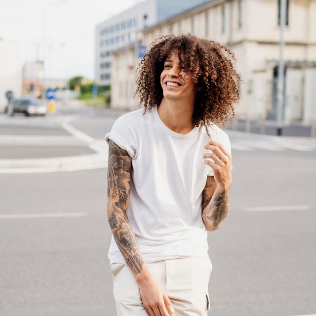smiling man with tattooed arms and long brown curly hair standing on a street