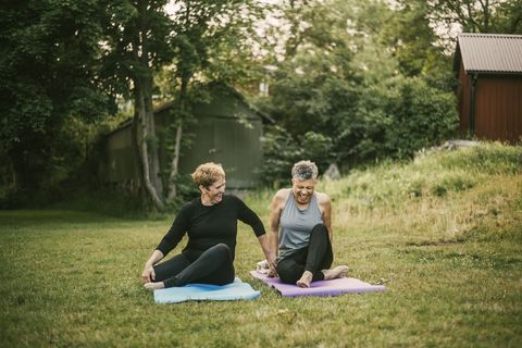 smiling woman assisting friend while exercising on mat in public park