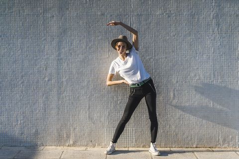Smiling woman wearing hat standing in front of tiled wall doing stretching exercises