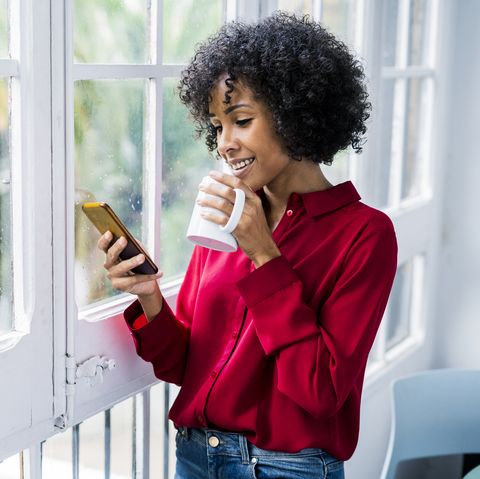 Smiling woman with cup of coffee and cell phone standing at the window at home