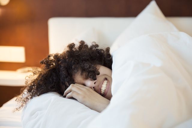 a smiling woman with dark curly hair in bed under the covers, head on a white pillow,