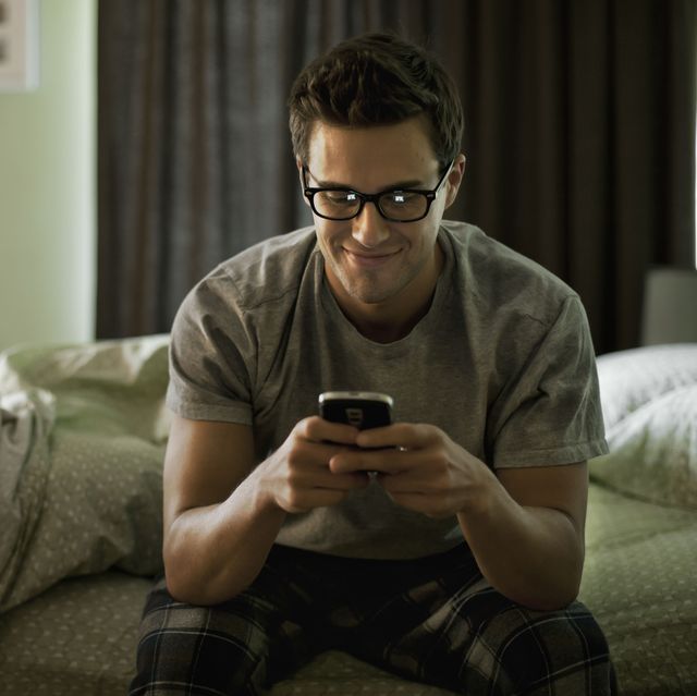 Smiling young man using phone while sitting on bed at home