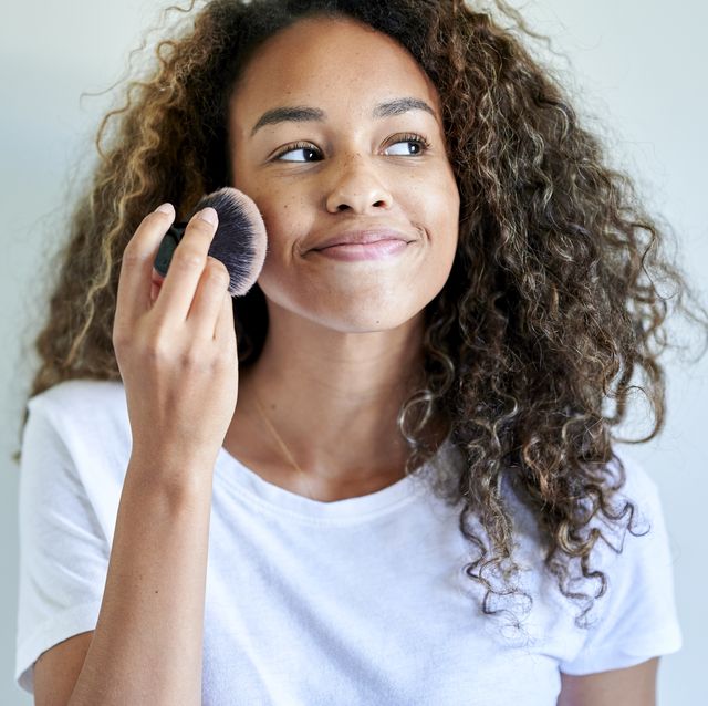 smiling young woman applying face powder with make up brush against white wall