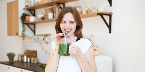 smiling young woman drinking green smoothie juice in kitchen