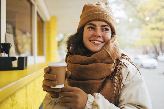 smiling young woman looking away with disposable coffee cup at street cafe during winter