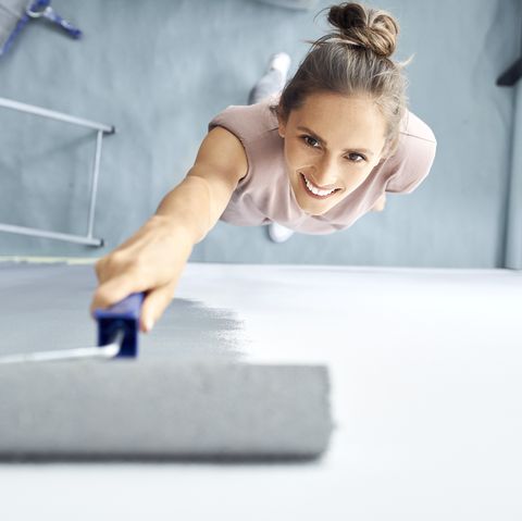 smiling young woman painting wall with paint roller while standing at home