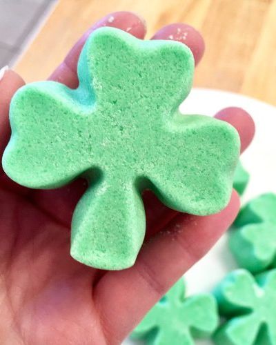 hand holding a green bath truffle shaped like a shamrock with more of the same shamrock bath truffles in the background