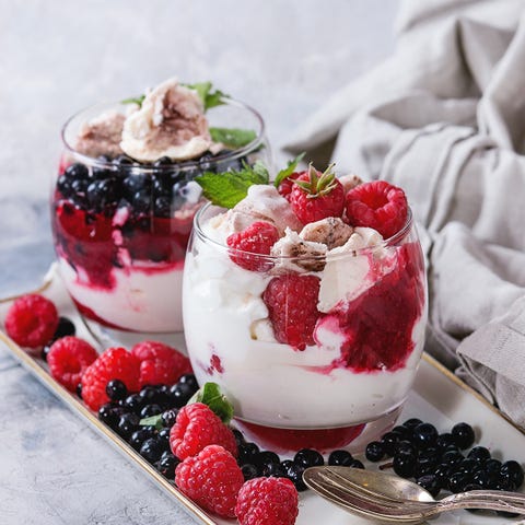 Traditional summer dessert Eton Mess. Broken meringue with whipped cream, berry jam, fresh blueberries and raspberries in two glasses, decorated with mint leaves over gray concrete background.