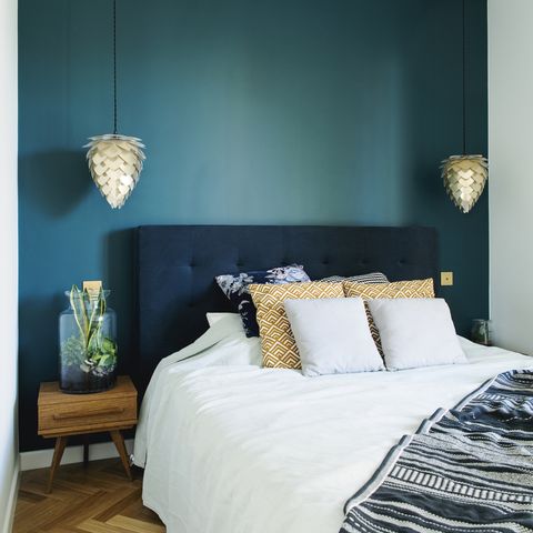 stylish bedroom interior with small wooden night table , garden in a jar, white bedding, colours pilows and blanket space with blue walls and brown wooden parquet design lamp