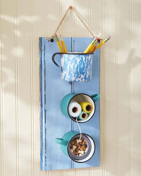 three enamelware mugs mounted on a piece of wood painted a summery blue serves as hanging storage piece for craft supplies, with pencils, spools of colorful thread, and push pins stashed in the mugs
