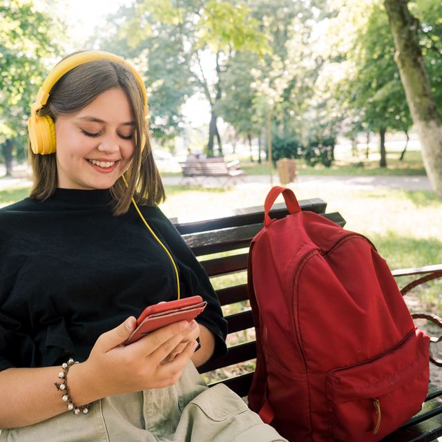 teen with yellow headphones sitting on bench with red backpack