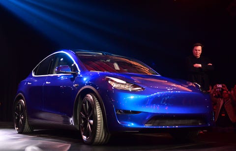 tesla model y on stage with elon musk in the background with arms folded staring at the camera