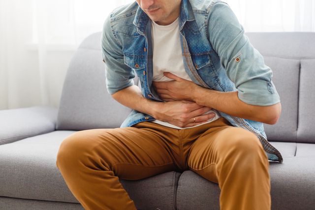 the man is sitting on a gray couch and holding his belly medicine and health concept, stomach problems the man suffers from stomach ache, gastric problems abdominal pain, suffering and pain