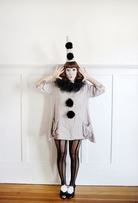woman dressed as a vintage clown with a gray peaked hat with black poms poms a long gray linen shirt with black pom poms black striped tights and black shoes with white pom poms her face is painted white with bowed red lips