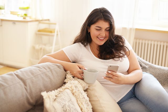 technology, communication and leisure concept pretty girl with chubby cheeks relaxing at home with electronic gadget overweight young brunette woman texting sms on smart phone and having coffee