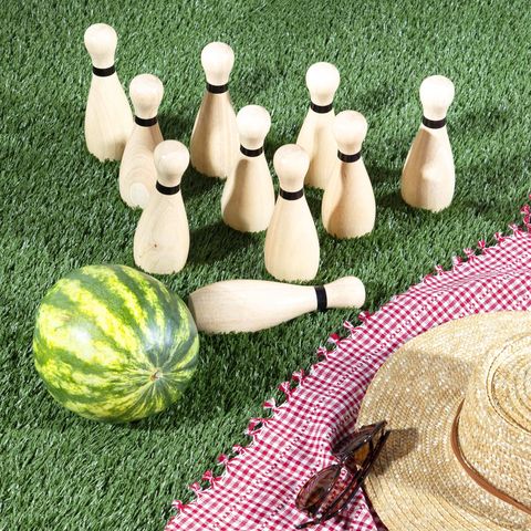 watermelon and bowling pins on the lawn next to a red and white gingham picnic blanket, straw hat, and sunglasses