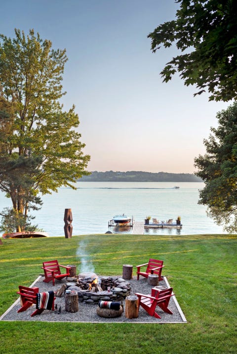 lakeside fire pit surrounded by 4 red adirondack chairs, log side tables, and blankets, overlooking evening sky