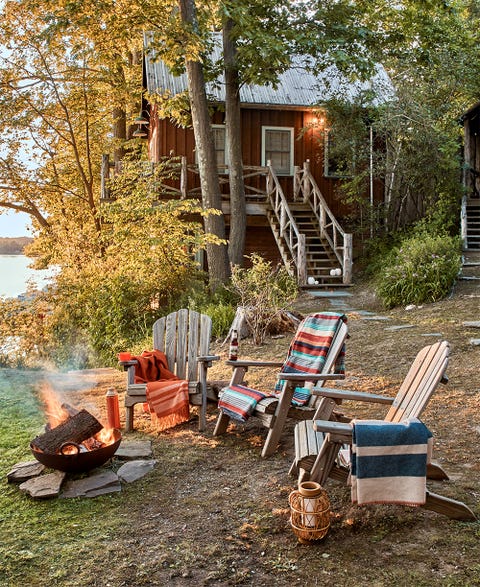 fire bowl surrounded by 3 adirondack chairs with blankets, rattan lantern outside lakeside cabin