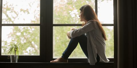 Thoughtful girl sitting on sill embracing knees looking at window