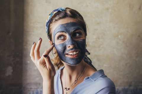 Thoughtful young woman smiling while applying facial mask against wall at home