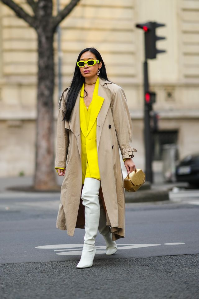 paris, france   march 02 miki cheung wears neon yellow sunglasses from versace, gold and rhinestones logo pendant earrings from valentino, gold necklace, a yellow silk v neck short dress, a matching yellow blazer jacket, a beige oversized long trench coat, a gold shiny leather handbag, silver and diamonds rings, white leather pointed  heels high boots  waders, gold rings, outside acne studios , during paris fashion week   womenswear fw 2022 2023, on march 02, 2022 in paris, france photo by edward berthelotgetty images