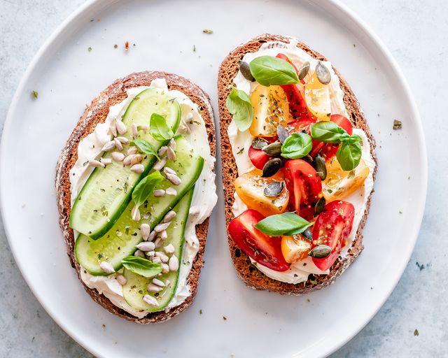two slices of toast with cream cheese, cucumber, tomato, pumpkin seeds, sesame seeds and basil