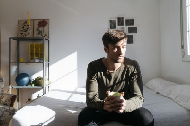 unhappy young man sitting on bed with cup of coffee