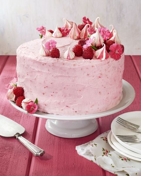 raspberry pink velvet cake with raspberry frosting on a white cake stand and garnished with fresh raspberries meringue cookies and small pink flowers