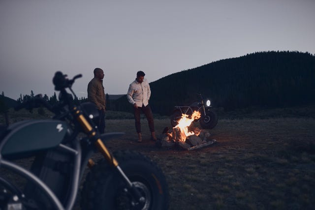 two men standing around a campfire at dusk with two volcon motorycycles
