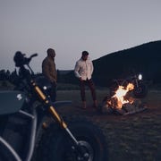 two men standing around a campfire at dusk with two volcon motorycycles