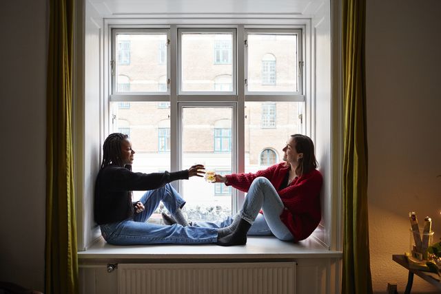 young female roommates toasting cocktails in apartment window during covid 19 isolation