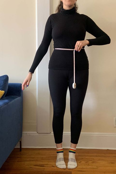 a woman wearing black leggings and a black turtleneck demonstrating how to measure your waist