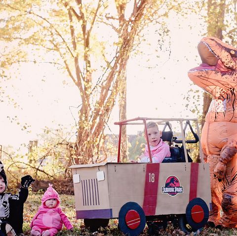 a family with jurassic park group halloween costumes including a baby in a dinosaur one piece costume a boy in a wheelchair adaptive jeep costume and an inflatable dinosaur costume