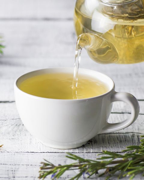 white cup of healthy rosemary tea pouring from teapot with fresh rosemary bunch on white wooden rustic background, winter herbal hot drink concept, salvia rosmarinus