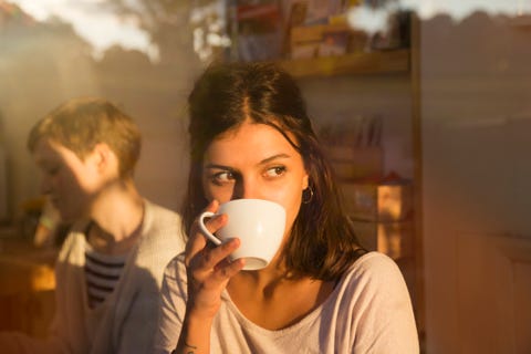 woman drinking coffee in urban cafe while looking outside, sunny reflections in window