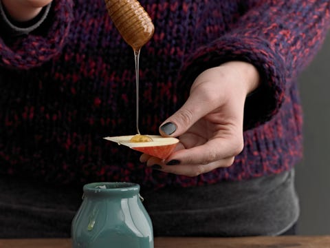 a woman drizzling honey from a round wooden drizzle stick on to an apple slice