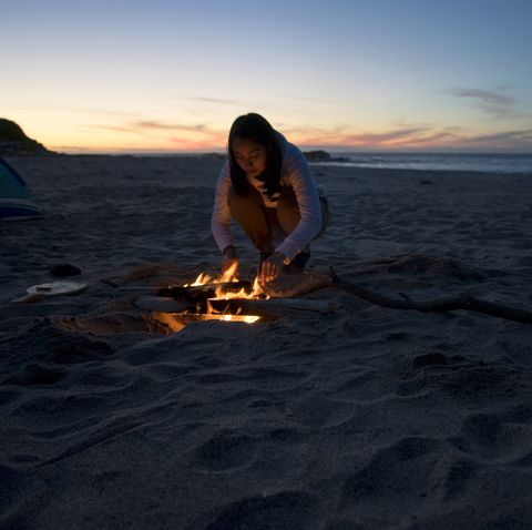 woman standing by campfire on beach against sky during sunset