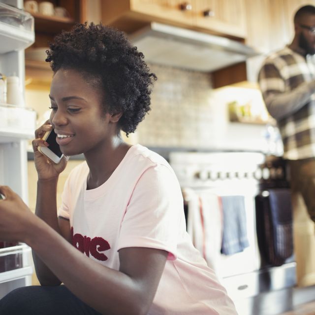Woman talking on smart phone, reading label on jar in refrigerator in kitchen