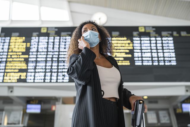 holiday travel  woman wearing protective face mask talking on smart phone at airport