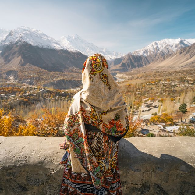 a woman wearing traditional dress sitting on wall and looking at hunza valley in autumn season, gilgit baltistan in pakistan