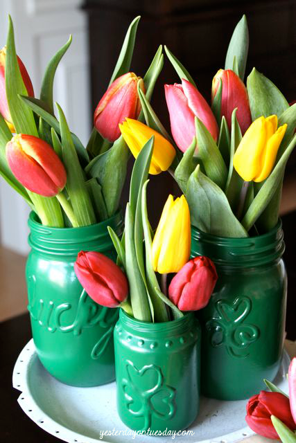 three mason jars painted green with puffy paint shamrocks on two of them and lucky written in puffy pain on the third all hold tulips