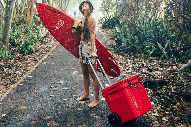 a woman holding a surfboard and rolling a red yeti cooler