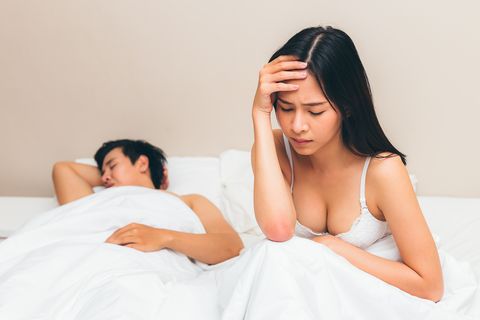 young asian couple having relationship problems with sex, depressed woman sitting on bed and her husband is sleeping at bedroom, failure sex concept