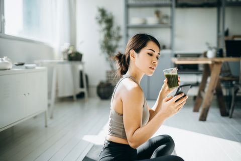 young asian sports woman resting and having green smoothie after exercising at home in the morning, using smartphone