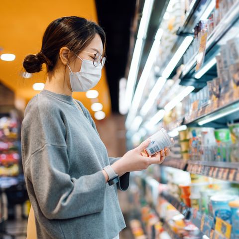 young asian woman grocery shopping in a supermarket standing by the refrigerated section, holding a pot of fresh organic healthy yoghurt reading the nutritional label and checking ingredients at the back