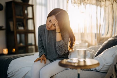 young asian woman sitting on the bed feeling sick and suffering from headache, a glass of water and medicine on the side table