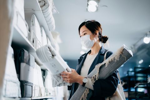 young asian woman with protective face mask shopping for stylish home decorations in a homeware store, carrying a carpet and choosing bedding sets
